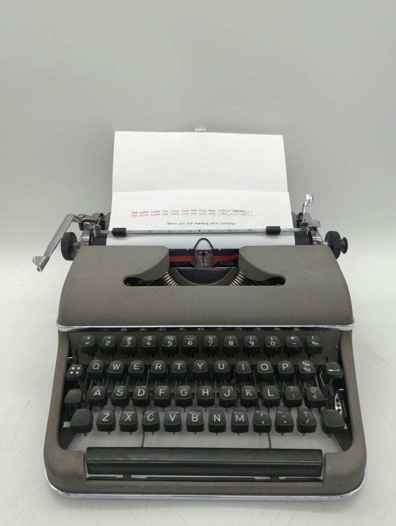 Pu&ograve; includere: Una macchina da scrivere vintage grigia con una tastiera nera e un foglio di carta nel carrello. Il testo sul foglio dice "The quick brown fox jumps over the lazy dog.  abcdefghijklmnopqrstuvwxyz!" e "Thank you for viewing this listing!"