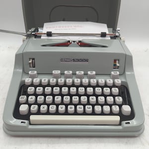 May include: A light gray Hermes 3000 typewriter with a QWERTY keyboard layout, white keys, and a paper carriage containing a sheet of paper with printed text. The machine features a metal body, a long space bar, and various function keys.