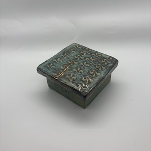 May include: A square ceramic box with a lid. The box is glazed in shades of teal and green, with a crack visible on the top. The lid features a repeating pattern of stylized floral designs.