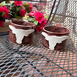 May include: Two ceramic mugs featuring a white longhorn steer design. The mugs are a deep red colour with brown speckles.