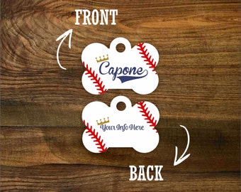 Puede incluir: Una etiqueta para perro de hueso blanco con un diseño de béisbol. El lado frontal presenta una corona dorada y el nombre "Capone" en una fuente cursiva azul marino. El lado posterior tiene una corona dorada y el texto "Your Info Here".