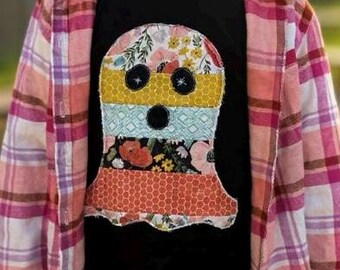 May include: A black t-shirt featuring a patchwork ghost design with floral and geometric patterns. The ghost has two black button eyes and a black button mouth. The shirt is worn with a pink and white plaid flannel shirt.
