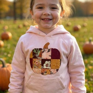 Op de afbeelding: Een lichtroze hoodie met een patchwork pompoenontwerp op de voorkant. De pompoen is gemaakt van verschillende stofpatronen en -kleuren, waaronder bloemen en effen vierkanten. Het kind lacht in een pompoenveld.