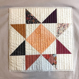 Puede incluir: Textil acolchado cuadrado con diseño de estrella. La estrella está compuesta por telas en tonos naranja, marrón, negro y burdeos, sobre un fondo crema. La pieza está cosida a máquina con un patrón vertical.