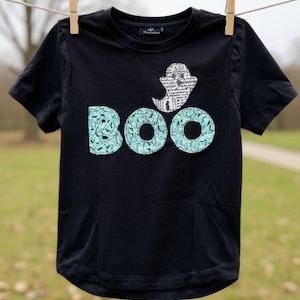 May include: Black t-shirt with a ghost and the word "BOO" in a teal font. The ghost is white with a newspaper print design. The letters "BOO" have a teal pattern. The shirt is hanging on a clothesline with wooden clothespins.