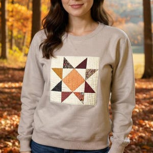 Könnte beinhalten: Beiges Sweatshirt mit einem gesteppten Stern-Design. Der Stern besteht aus verschiedenen Stoffmustern in Orange-, Burgunder-, Schwarz- und gemusterten Farbtönen, vor einem cremefarbenen Hintergrund. Das Sweatshirt hat einen Rundhalsausschnitt und lange Ärmel.