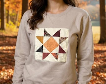 May include: A tan sweatshirt featuring a quilted star design. The star is composed of various fabric patterns in shades of orange, burgundy, black, and patterned fabrics, set against a cream-colored background. The sweatshirt has a crew neck and long sleeves.