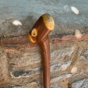 May include: A handcrafted wooden walking stick with a rounded, light-colored top and a dark brown shaft. The handle has a unique, carved design. The stick is leaning against a stone wall.