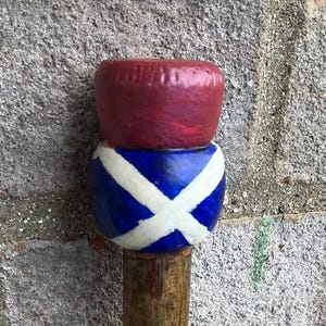 May include: A wooden walking stick featuring a decorative topper. The topper consists of a dark red cylindrical base topped with a blue sphere painted with a white saltire cross pattern. The shaft is made of natural brown wood with a textured surface.