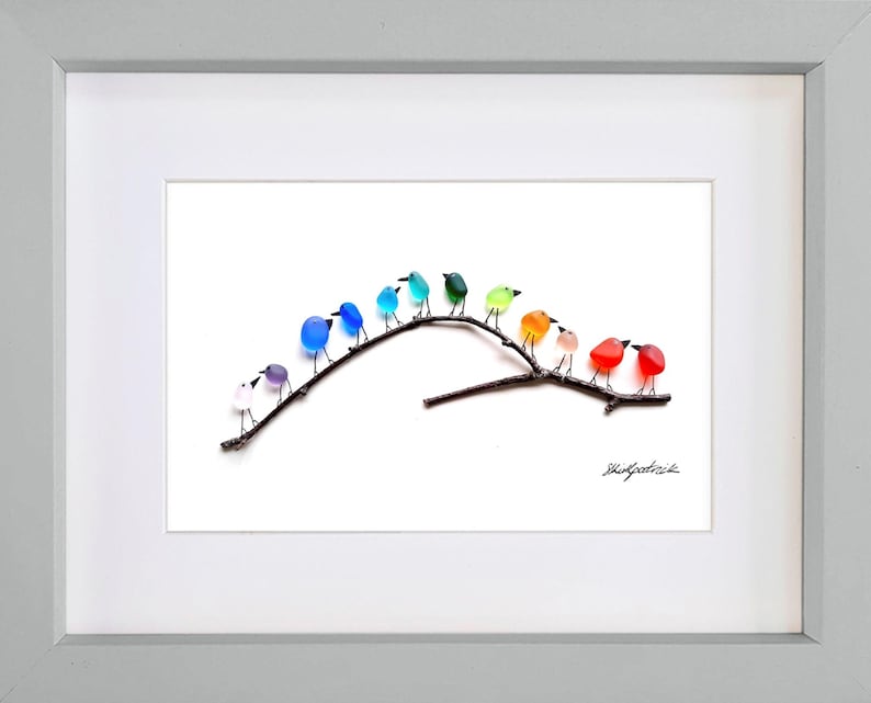 May include: A framed artwork featuring a collection of colorful glass birds perched on a dark brown branch. The birds are in various colors, including blue, green, orange, and red. The artwork is set against a white background and framed in a light gray frame.