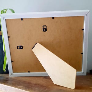 May include: A white picture frame with a light brown backing and a beige easel stand. The frame is resting on a wooden surface, with a green plant in a blue pot and purple flowers in the background.