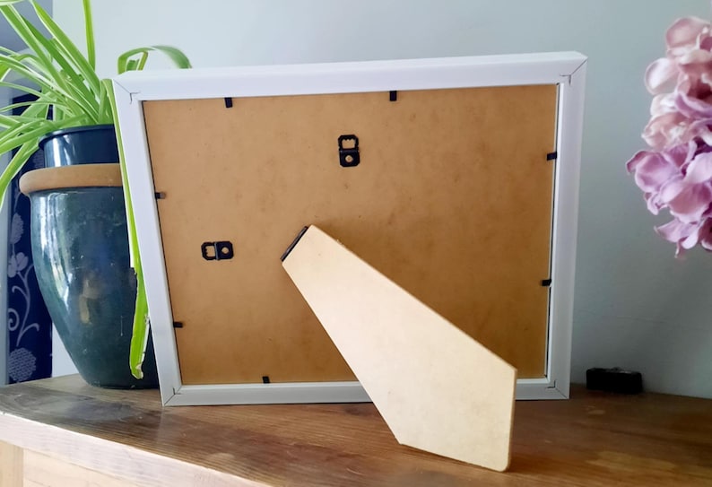 May include: A white picture frame with a light brown backing and a beige easel stand. The frame is resting on a wooden surface, with a green plant in a blue pot and purple flowers in the background.