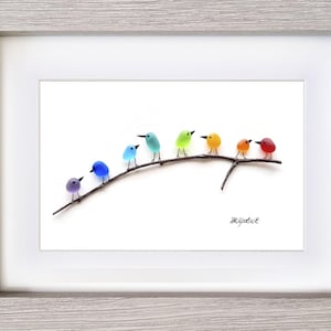 May include: A framed artwork featuring a row of colorful glass birds perched on a dark branch against a white background. The birds are in shades of purple, blue, green, yellow, orange, and red. The frame is light gray.