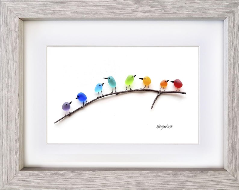 May include: A framed artwork featuring a row of colorful glass birds perched on a dark branch against a white background. The birds are in shades of purple, blue, green, yellow, orange, and red. The frame is light gray.