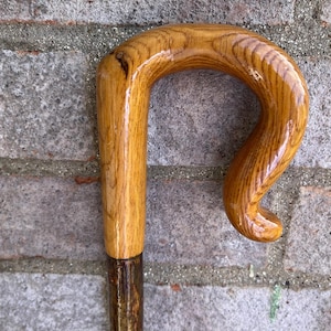 Pode incluir: Uma bengala com um cabo de madeira castanho-claro polido, com uma forma curva e ergonómica e um padrão de grão de madeira visível. O cabo está fixado a um eixo de madeira mais escuro e texturado, apresentando um acabamento natural.