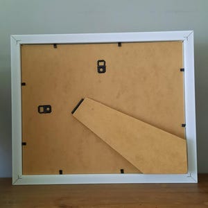 May include: A white square picture frame, viewed from the back, with a brown cardboard backing and a built-in easel. The frame has a simple, clean design, suitable for displaying photos or artwork. The frame is resting on a wooden surface.