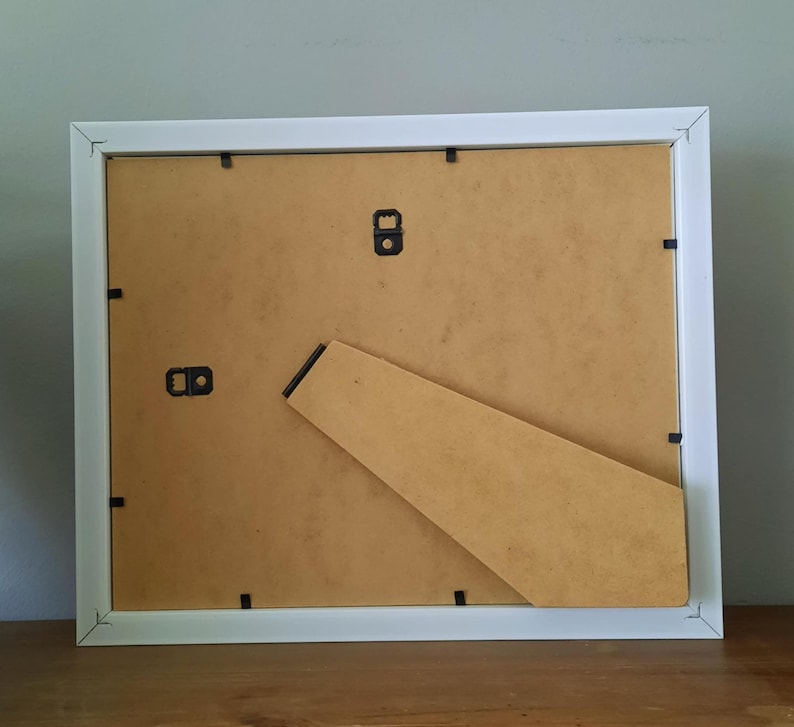 May include: A white square picture frame, viewed from the back, with a brown cardboard backing and a built-in easel. The frame has a simple, clean design, suitable for displaying photos or artwork. The frame is resting on a wooden surface.