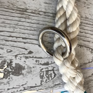 May include: A close-up of a white cotton rope with a silver metal ring attached. The rope is braided and the ring is smooth and shiny.