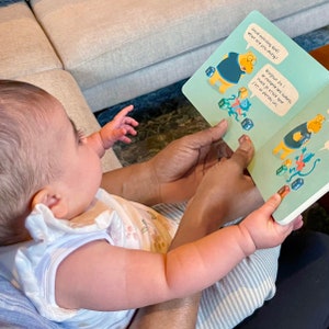 May include: A baby is sitting on a person's lap and looking at a book with colourful illustrations. The book has the text "Good morning No&eacute;! What are you doing?" and "Bonjour Zo! Je compte les cubes, mais je crois que j'en ai perdu un."