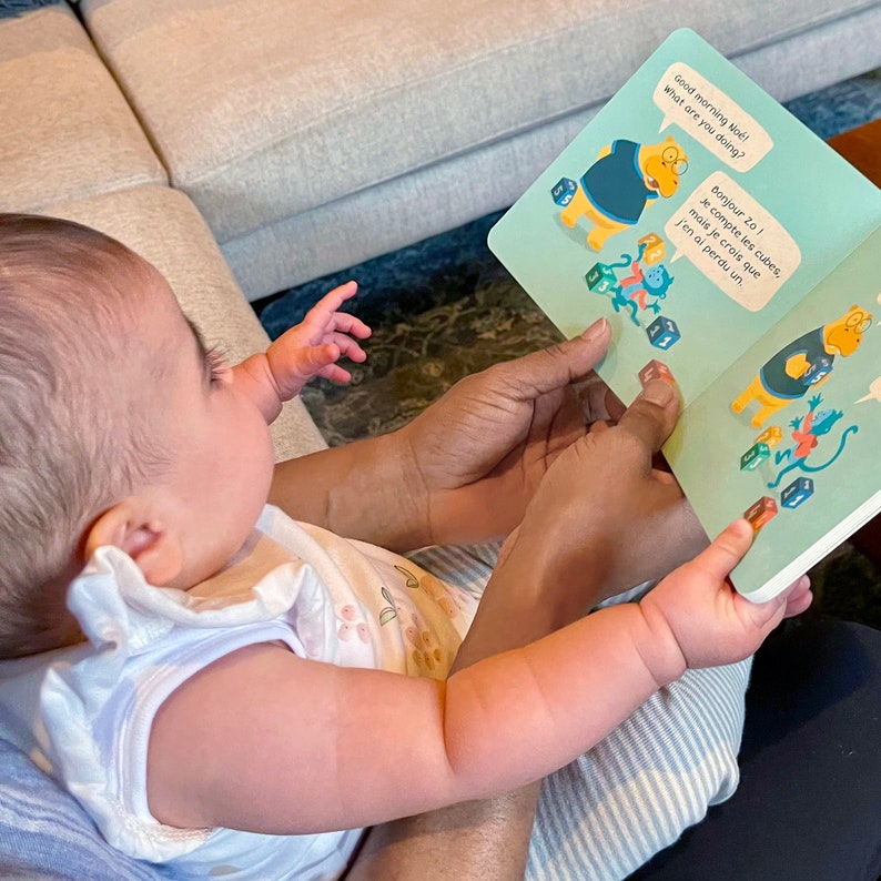 May include: A baby is sitting on a person's lap and looking at a book with colourful illustrations. The book has the text "Good morning No&eacute;! What are you doing?" and "Bonjour Zo! Je compte les cubes, mais je crois que j'en ai perdu un."