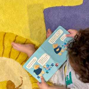 May include: A child is reading a book with colourful illustrations of a blue and white character counting cubes. The book has the text "Good morning Noel! What are you doing?" and "Bonjour Ze! Je compte les cubes, mais je crois que j'en ai perdu un."  The text "I'm sorry! Sometimes I get all confused too. Is this what you're looking for?" and "Oh oui! Merci 20" are also visible.