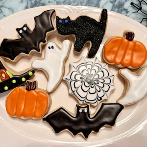 May include: A collection of Halloween-themed sugar cookies on a white oval platter. The cookies include two black bats, two white ghosts, two orange pumpkins, a black cat with textured sugar, a witch hat, and a circular spider web cookie.
