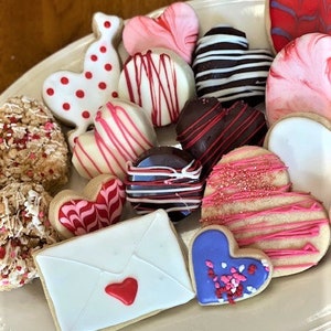 May include: A variety of Valentine's Day cookies decorated with red, pink, white, and blue icing. The cookies are shaped like hearts, envelopes, and other designs. Some cookies are topped with sprinkles.