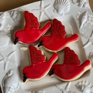 May include: Four red cardinal bird-shaped sugar cookies decorated with red icing and red sprinkles on a white plate with a seashell design.