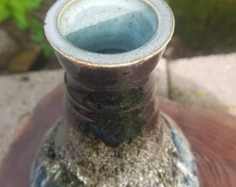 May include: A handcrafted ceramic vase with a wide, flared rim and a bulbous body. The vase features a dark brown neck and a base with blue, green, and white glaze. The vase is approximately 7 inches tall.