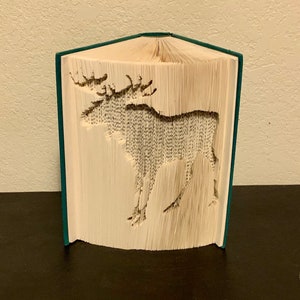 Peut inclure: Une sculpture d'art de livre représentant un élan, créée en pliant les pages d'un livre. Le livre est relié d'une couverture vert foncé.