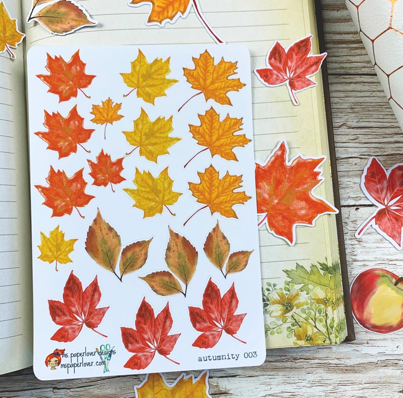 Puede incluir: Una hoja de pegatinas de hojas de oto&ntilde;o coloridas con una variedad de formas y tama&ntilde;os. Las pegatinas est&aacute;n sobre un fondo blanco y presentan tonos de rojo, naranja, amarillo y marr&oacute;n. El texto "autumunity 003" est&aacute; impreso en la esquina inferior derecha.
