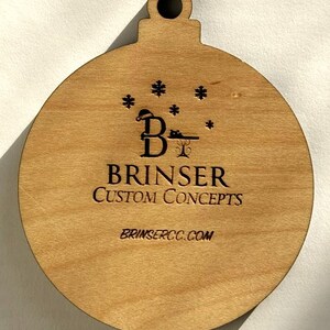 Puede incluir: Adorno navide&ntilde;o de madera en forma de bola con un lazo en la parte superior. El adorno tiene grabado el texto "BRINSER CUSTOM CONCEPTS" y "BRINSERCC.COM", junto con un logotipo con una "B" y copos de nieve.