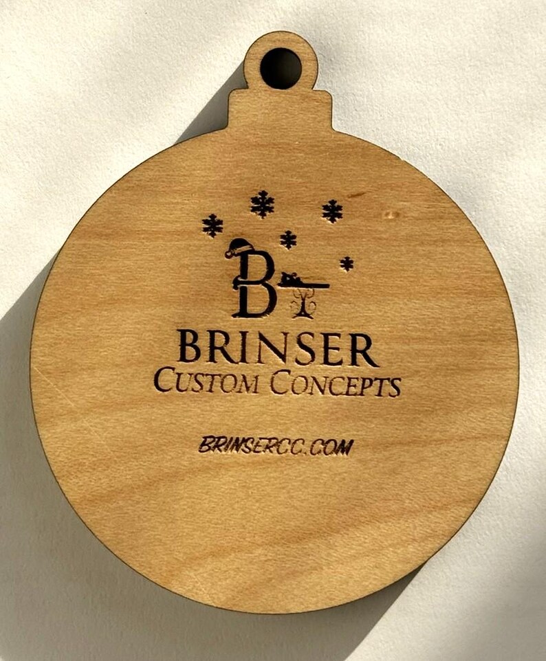 Puede incluir: Adorno navide&ntilde;o de madera en forma de bola con un lazo en la parte superior. El adorno tiene grabado el texto "BRINSER CUSTOM CONCEPTS" y "BRINSERCC.COM", junto con un logotipo con una "B" y copos de nieve.