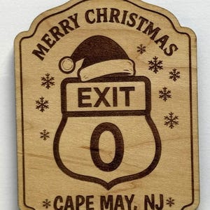 May include: Wooden Christmas ornament with the words "MERRY CHRISTMAS" and "EXIT 0 CAPE MAY, NJ" engraved. The ornament features a Santa hat above the exit sign and snowflakes. The ornament has a hole at the top for hanging.