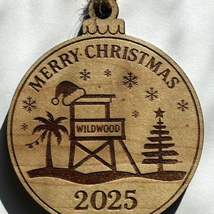 Puede incluir: Adorno navide&ntilde;o redondo de madera con tem&aacute;tica playera. El adorno presenta las palabras "MERRY CHRISTMAS" y "WILDWOOD", un puesto de salvavidas, una palmera, un &aacute;rbol de Navidad, copos de nieve y el a&ntilde;o "2025". Tiene una cuerda de yute para colgar.