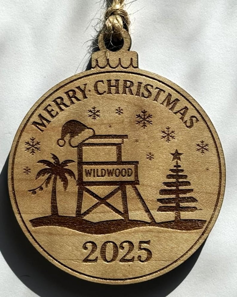 Puede incluir: Adorno navide&ntilde;o redondo de madera con tem&aacute;tica playera. El adorno presenta las palabras "MERRY CHRISTMAS" y "WILDWOOD", un puesto de salvavidas, una palmera, un &aacute;rbol de Navidad, copos de nieve y el a&ntilde;o "2025". Tiene una cuerda de yute para colgar.