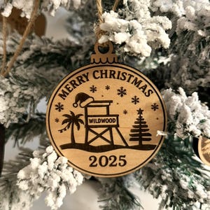 Puede incluir: Adorno navide&ntilde;o circular de madera con la inscripci&oacute;n "Merry Christmas" y el a&ntilde;o "2025". El dise&ntilde;o incluye un puesto de salvavidas, una palmera, un &aacute;rbol de Navidad, copos de nieve y un gorro de Pap&aacute; Noel.