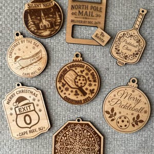 Puede incluir: Una colecci&oacute;n de adornos navide&ntilde;os de madera cortados con l&aacute;ser. Los dise&ntilde;os incluyen una escena de iglesia, un barco, una se&ntilde;al de tr&aacute;fico, una paleta de pickleball y un copo de nieve. Cada adorno tiene un agujero para colgar y presenta texto grabado.