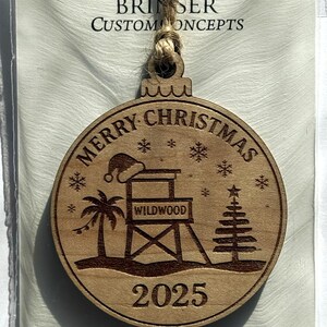 Puede incluir: Adorno navide&ntilde;o de madera grabado con l&aacute;ser con tem&aacute;tica playera. El adorno presenta las palabras "Merry Christmas" y "Wildwood 2025" junto con un puesto de salvavidas, una palmera y un &aacute;rbol de Navidad. Incluye un colgador de cuerda de yute.