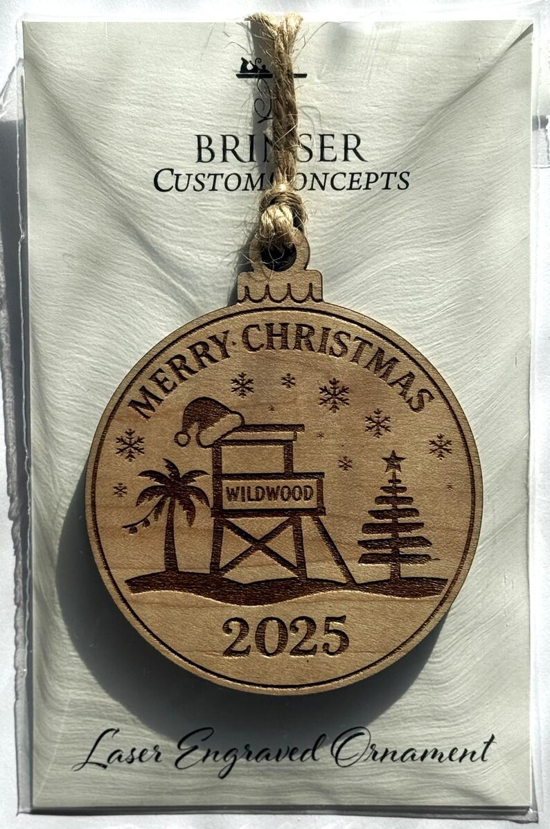 Puede incluir: Adorno navide&ntilde;o de madera grabado con l&aacute;ser con tem&aacute;tica playera. El adorno presenta las palabras "Merry Christmas" y "Wildwood 2025" junto con un puesto de salvavidas, una palmera y un &aacute;rbol de Navidad. Incluye un colgador de cuerda de yute.