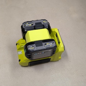K&ouml;nnte beinhalten: Ein gelbes und schwarzes Ryobi 40V Lithium-Ionen-Batterieladeger&auml;t mit zwei &uuml;bereinander gestapelten Batterien. Die Batterien sind mit "Ryobi ONE+ EDGE 4.0Ah" beschriftet.