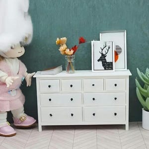 May include: White chest of drawers with nine drawers and black knobs. The dresser is on a wooden floor with a chevron pattern. There are two framed pictures above the dresser, one with a deer silhouette and the other with a geometric design. A vase with dried flowers is on the dresser.