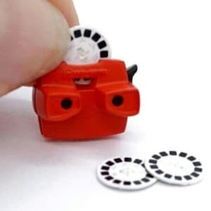Puede incluir: Un juguete view-master de plástico rojo con dos agujeros de visión redondos y una ranura para un carrete de película. Tres carretes de película de plástico blanco con imágenes negras están sobre una superficie blanca.