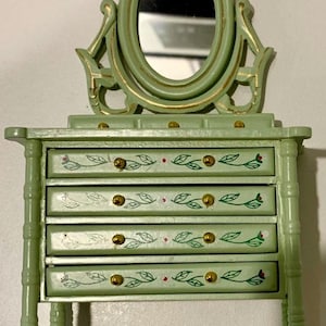 May include: A miniature green vanity unit with an oval mirror and four drawers. The drawers feature leaf and berry designs and gold-coloured knobs. The mirror and frame have gold accents. The vanity is a decorative item.