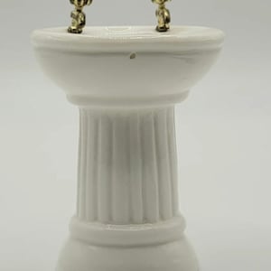 May include: A miniature white porcelain pedestal sink with two small gold-colored figures on the rim. The sink has a fluted column base and a rounded basin, perfect for dollhouses or miniature displays.