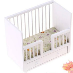 May include: A miniature white wooden crib with a floral patterned mattress. The crib has vertical slats on the sides and a drawer at the bottom. The mattress features a pattern of green leaves and purple flowers.