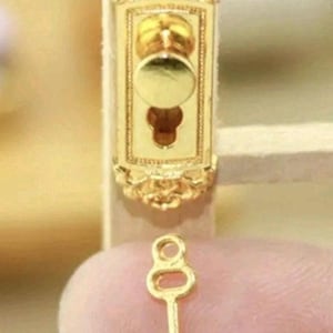 Peut inclure: Une plaque de porte miniature en métal doré présentant un cadre rectangulaire orné avec un bouton rond central et une serrure, accompagnée d'une petite clé en métal doré assortie avec un anneau circulaire et un panneton simple.