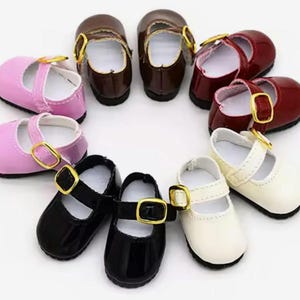 Puede incluir: Conjunto de diez zapatos de muñeca en varios colores, incluyendo rosa, blanco, negro, marrón y rojo. Los zapatos tienen un estilo Mary Jane con una correa sobre el empeine y un cierre de hebilla.