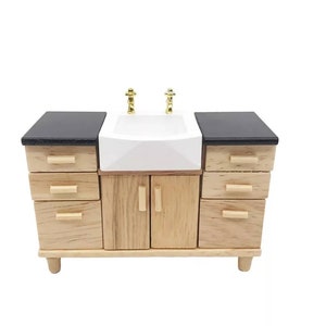 May include: A miniature wooden kitchen sink cabinet with a white sink, gold taps, and a black countertop. The cabinet has multiple drawers and doors, and is made of light-coloured wood.