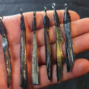 Op de afbeelding: Een verzameling van zes zwarte kyaniet hangers met zilverkleurige draad lussen. De kyaniet bladen variëren in grootte en hebben een natuurlijke, getextureerde uitstraling. De hangers worden in een hand gehouden, wat hun unieke vormen en kleuren laat zien.